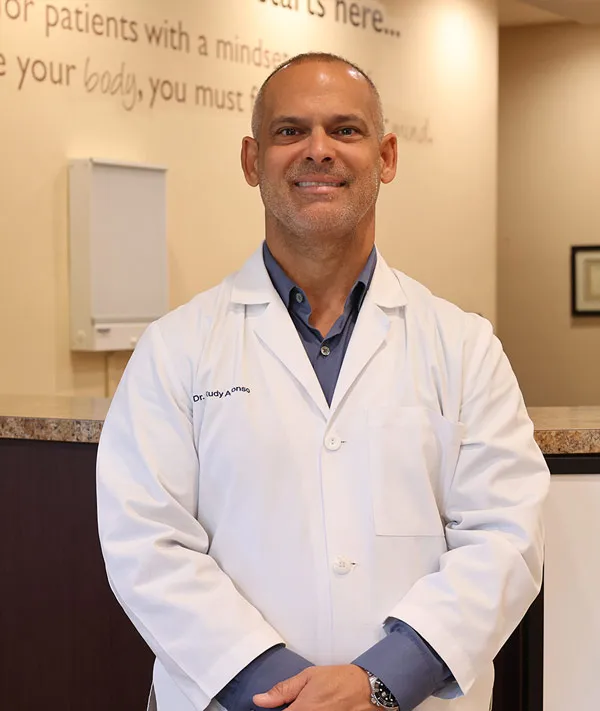 Dr. Rodolfo Alfonso standing in the clinic lobby wearing a white lab coat and smiling confidently.