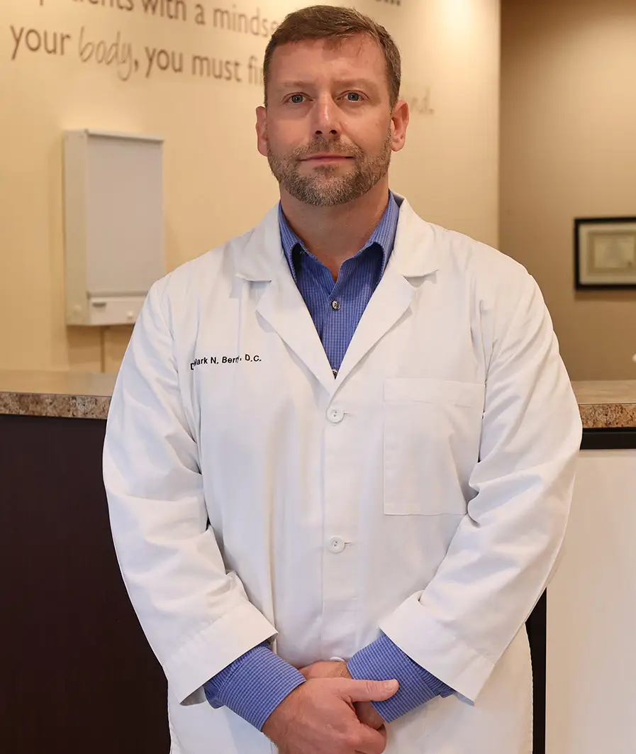 Doctor wearing a white coat and blue shirt, standing seriously in a clinic in front of a counter.