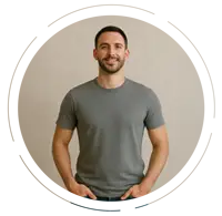 Confident young man standing tall with hands in pockets, smiling, wearing a gray T-shirt and blue jeans against a beige background.